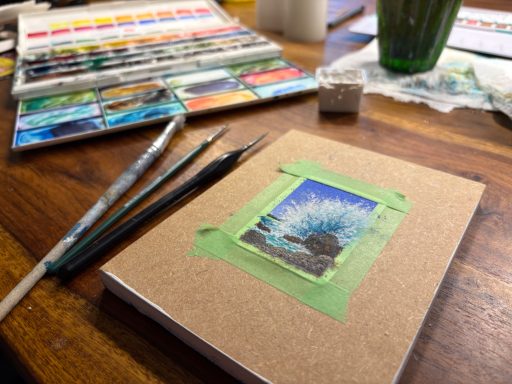 Ein Arbeitsplatz mit Aquarellfarben, Pinseln und einem Notizbuch mit festgeklebtem Bild von spritzender Meeresgischt